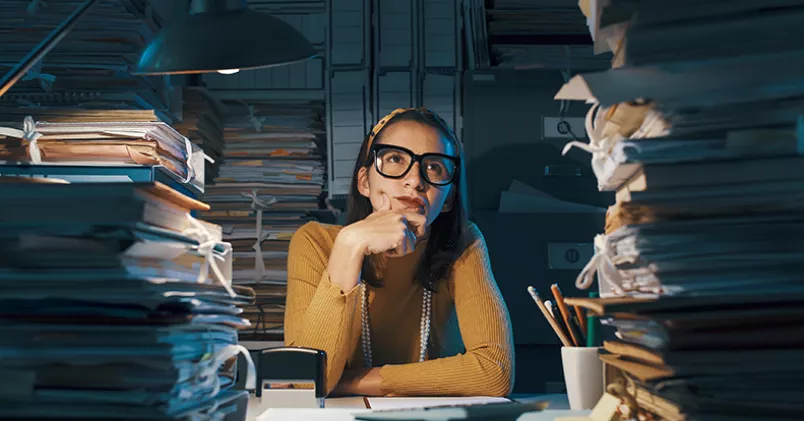 Kvinna vid skrivbord med höga pappershögar.