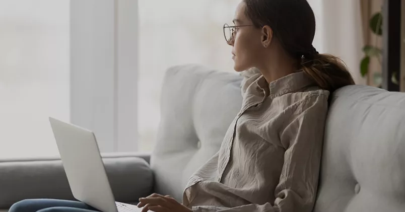 Nära var tredje anställd under 30 år är orolig över att bli uppsagd.  Kvinna sitter i soffan med laptop i knät och tittar ut genom fönstret.