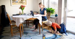 Kvinna vid köksbord med bärbar dator. Hon arbetar. På golvet ett barn som är hemma och leker samtidigt som mamman arbetar. 