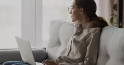 Kvinna sitter i soffan med laptop i knät och tittar ut genom fönstret.